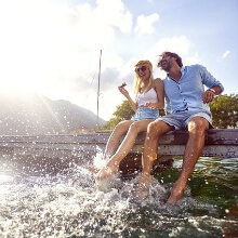 Two people splash around on the jetty in front of a mountain backdrop, one is holding an ice cream – summer joy in the travel worlds eMagazine 2025.