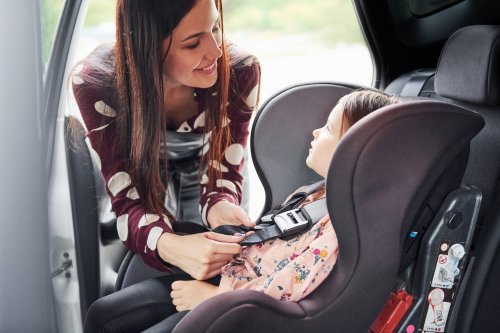 Mother securing her young daughter in a car seat next to a window fitted with Solarplexius sun protection