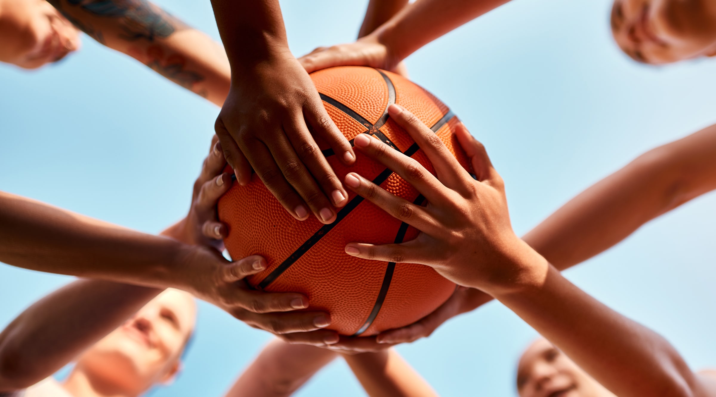 Group of hands reaching together for a basketball against a blue sky